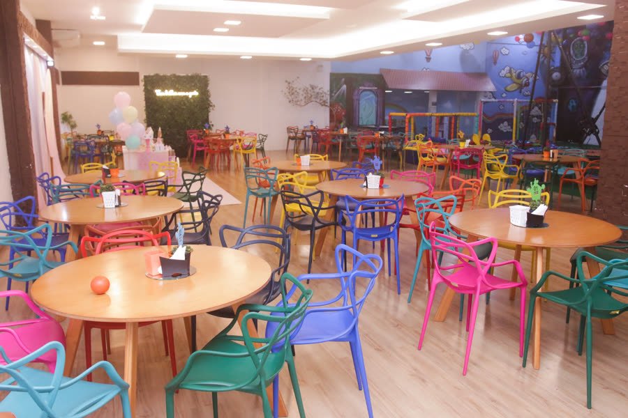 Salão integrado com mesas coloridas e área de brinquedos ao fundo